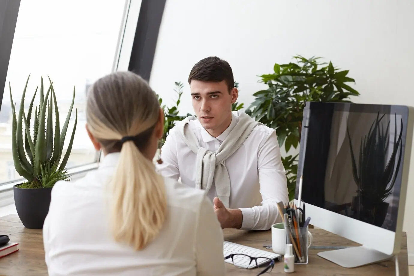 A handsome, serious young male HR specialist interviews an unrecognizable woman with a ponytail during a job interview, asking about her experience and skills. Employment and human resources.